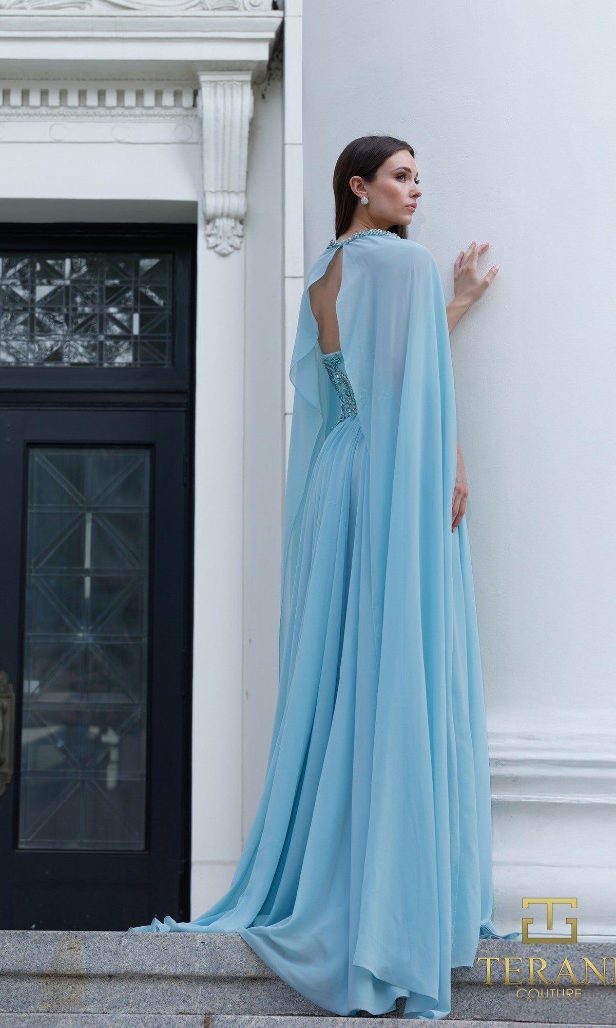 A woman in the Terani 261M6402 long formal dress, featuring a light blue hue, embellished bodice, cape detail, and lace accents, stands on stone steps before a white columned building.